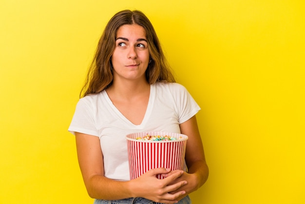 Junge Kaukasierin, die ein Popcorn isoliert auf gelbem Hintergrund hält, verwirrt, fühlt sich zweifelhaft und unsicher.