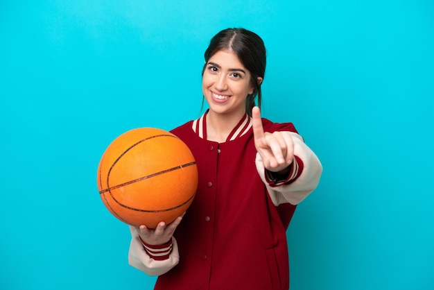 Junge kaukasische Basketballspielerin isoliert auf blauem Hintergrund, die einen Finger zeigt und hebt