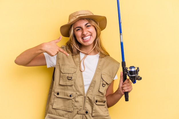 Junge kaukasische blonde Fischerin, die eine Stange einzeln auf gelbem Hintergrund hält, die mit der Hand auf einen Hemdkopierraum zeigt, stolz und selbstbewusst