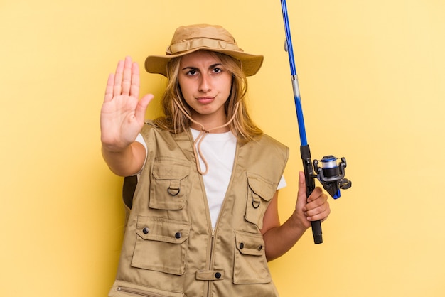 Junge kaukasische blonde Fischerin, die eine Stange isoliert auf gelbem Hintergrund hält, die mit ausgestreckter Hand steht und ein Stoppschild zeigt, um Sie zu verhindern.