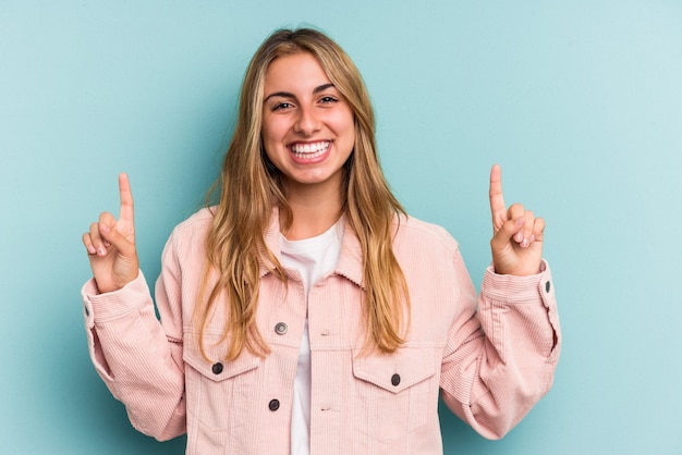 Junge kaukasische blonde Frau, die auf blauem Hintergrund isoliert ist, zeigt an, dass beide Vorderfinger eine Leerstelle zeigen.