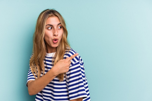 Junge kaukasische blonde Frau, die auf blauem Hintergrund isoliert ist, zeigt mit dem Daumenfinger weg, lacht und sorglos.