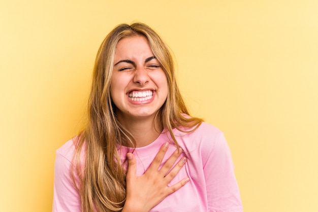 Junge kaukasische blonde Frau, die auf gelbem Hintergrund isoliert ist, lacht laut und hält die Hand auf der Brust.