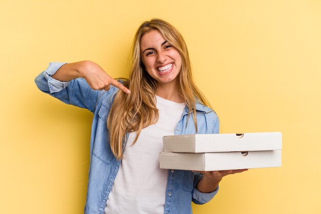 Junge kaukasische blonde Frau, die Pizzas einzeln auf gelbem Hintergrund hält, die mit der Hand auf einen Hemdkopierraum zeigt, stolz und selbstbewusst