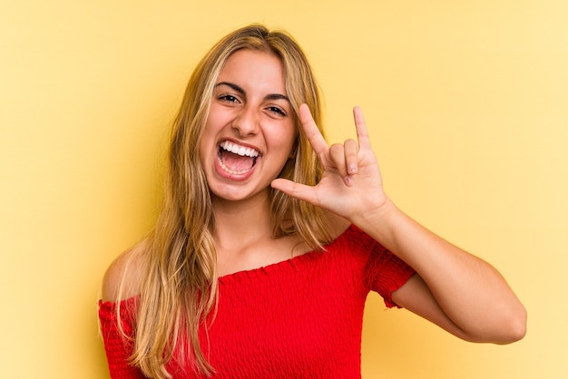 Junge kaukasische blonde Frau isoliert auf gelbem Hintergrund, die eine Hörnergeste als Revolutionskonzept zeigt.