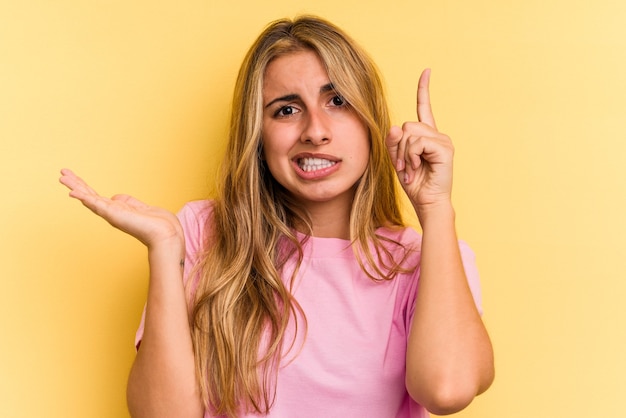 Junge kaukasische blonde Frau lokalisiert auf gelbem Hintergrund, der ein Produkt an Hand hält und zeigt.