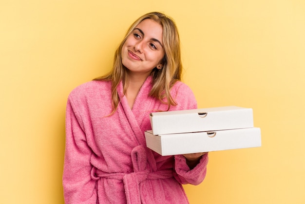 Junge kaukasische blonde Frau mit Bademantel, die Pizzas isoliert auf gelbem Hintergrund hält und davon träumt, Ziele und Zwecke zu erreichen