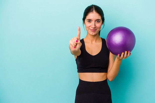 Junge kaukasische Eignungsfrau, die einen Ball lokalisiert auf blauem Hintergrund hält, der Nummer eins mit dem Finger zeigt.