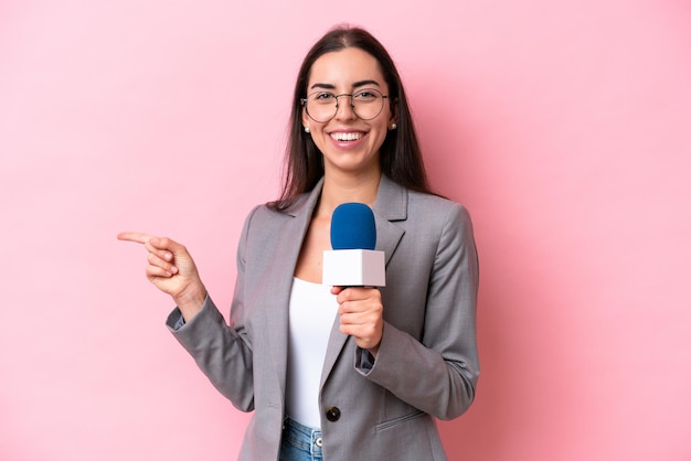 Junge kaukasische Fernsehmoderatorin isoliert auf rosafarbenem Hintergrund, die mit dem Finger zur Seite zeigt