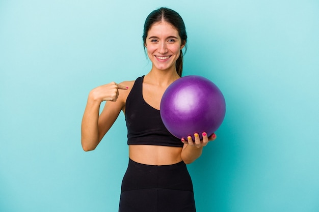 Foto junge kaukasische fitnessfrau, die einen ball auf blauem hintergrund hält, der mit der hand auf einen hemdkopierraum zeigt, stolz und selbstbewusst