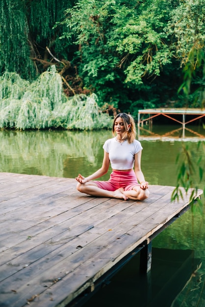 Junge kaukasische Fitnessfrau, die Yoga macht, meditiert im Freien, auf dem hölzernen Piersee am Sommertag.