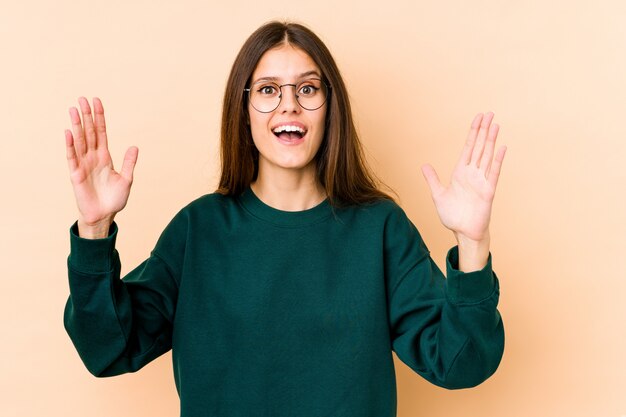 Junge kaukasische Frau auf beige Wand, die eine angenehme Überraschung empfängt, aufgeregt und Hände erhebt.