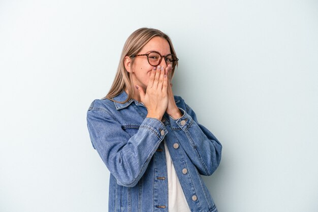 Junge kaukasische Frau auf blauem Hintergrund isoliert, die über etwas lacht und den Mund mit den Händen bedeckt.