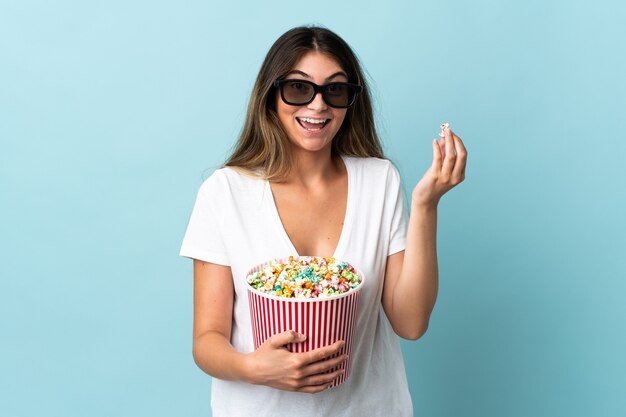 Junge kaukasische Frau auf blauem Hintergrund mit 3D-Brille isoliert und hält einen großen Eimer Popcorn