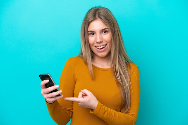 Junge kaukasische Frau auf blauem Hintergrund mit Handy isoliert und darauf zeigend