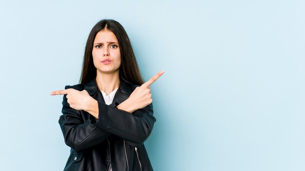 Junge kaukasische Frau auf blauer Wand zeigt seitwärts, versucht, zwischen zwei Optionen zu wählen.