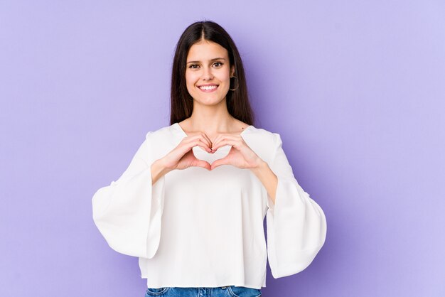 Junge kaukasische Frau auf lila Wand lächelnd und zeigt eine Herzform mit Händen.