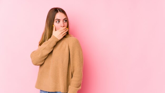 Junge kaukasische Frau auf rosa Wand nachdenklich schauend zu einem leeren Raum, der Mund mit Hand bedeckt.