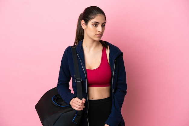 Junge kaukasische Frau des Sports mit Sporttasche lokalisiert auf rosa Hintergrund, der zur Seite schaut
