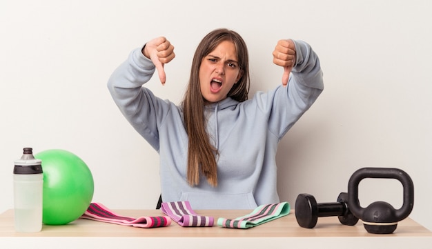 Foto junge kaukasische frau, die an einem tisch mit sportausrüstung sitzt, die auf weißem hintergrund lokalisiert wird, der daumen nach unten zeigt und abneigung ausdrückt.