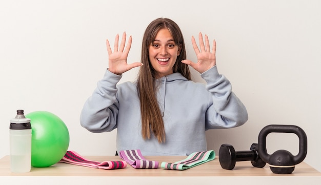 Junge kaukasische Frau, die an einem Tisch mit Sportausrüstung sitzt, die auf weißem Hintergrund lokalisiert wird, der Nummer zehn mit den Händen zeigt.