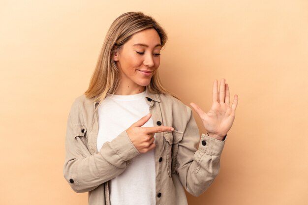 Junge kaukasische Frau, die auf beigefarbenem Hintergrund isoliert ist, lächelt fröhlich und zeigt Nummer fünf mit den Fingern.