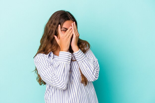 Junge kaukasische Frau, die auf blauem Hintergrund isoliert ist, blinzelt durch die Finger in die Kamera, verlegen das Gesicht.