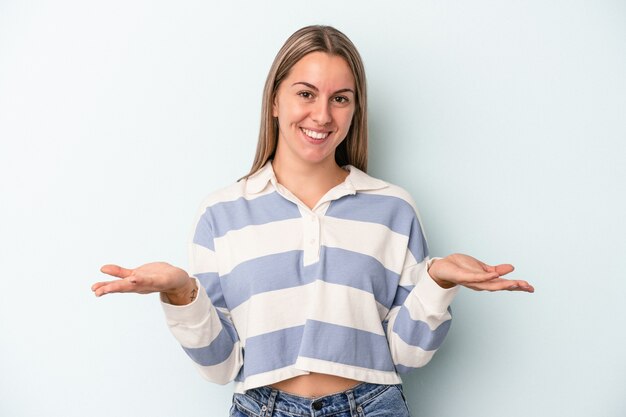 Junge kaukasische Frau, die auf blauem Hintergrund isoliert ist, macht mit Armen eine Waage, fühlt sich glücklich und selbstbewusst.