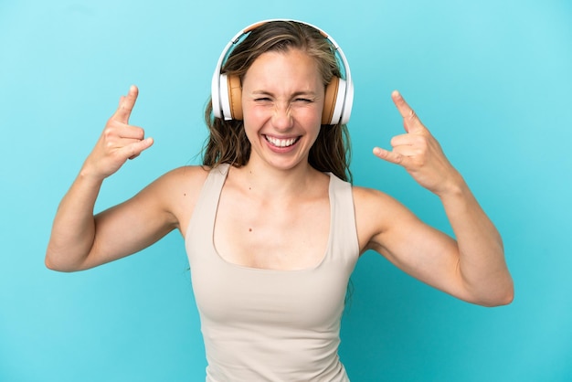 Junge kaukasische Frau, die auf blauem Hintergrund isoliert ist und Musik hört, die Rockgeste macht?