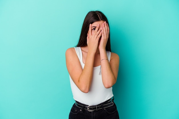 Junge kaukasische Frau, die auf blauer Wand isoliert ist, blinzelt durch erschrockene und nervöse Finger.