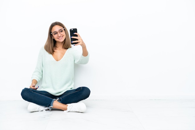 Junge kaukasische Frau, die auf dem Boden lokalisiert auf weißem Hintergrund sitzt und ein selfie macht