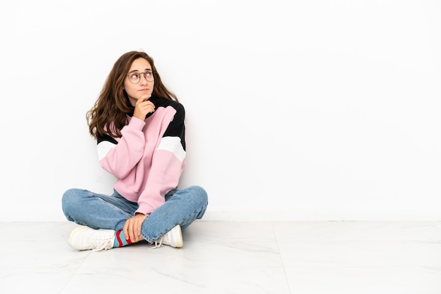 Junge kaukasische Frau, die auf dem Boden sitzt, isoliert auf weißem Hintergrund, hat Zweifel und einen verwirrten Gesichtsausdruck