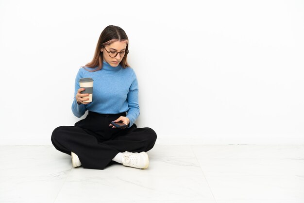 Junge kaukasische Frau, die auf dem Boden sitzt und Kaffee hält, um und ein Handy wegzunehmen