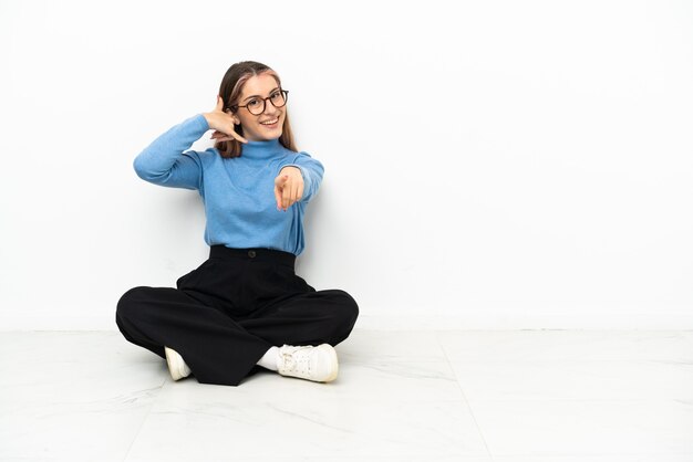 Junge kaukasische Frau, die auf dem Boden sitzt und Telefongeste macht und nach vorne zeigt