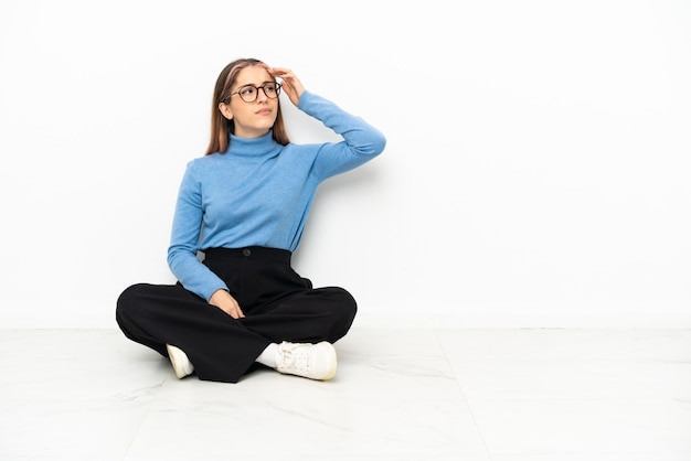 Junge kaukasische Frau, die auf dem Boden sitzt und Zweifel und mit verwirrendem Gesichtsausdruck hat