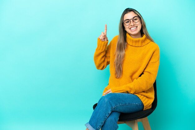 Junge kaukasische Frau, die auf einem Stuhl lokalisiert auf blauem Hintergrund sitzt und einen Finger im Zeichen des Besten zeigt