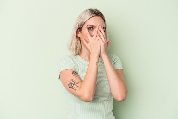 Junge kaukasische Frau, die auf grünem Hintergrund isoliert ist, blinzelt erschrocken und nervös durch die Finger.