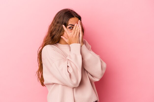 Junge kaukasische Frau, die auf rosafarbenem Hintergrund isoliert ist, blinzelt erschrocken und nervös durch die Finger.