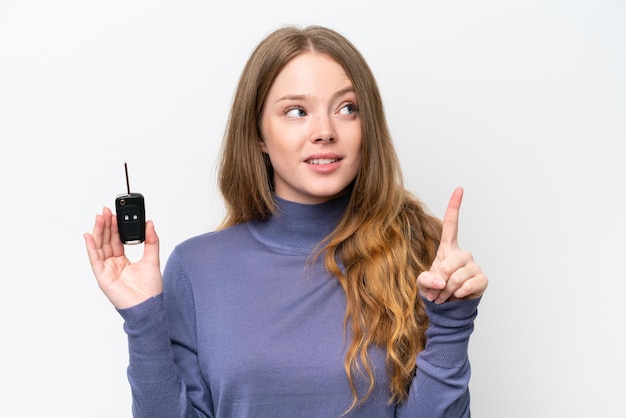 Junge kaukasische Frau, die Autoschlüssel isoliert auf weißem Hintergrund hält und beabsichtigt, die Lösung zu realisieren, während sie einen Finger hochhebt