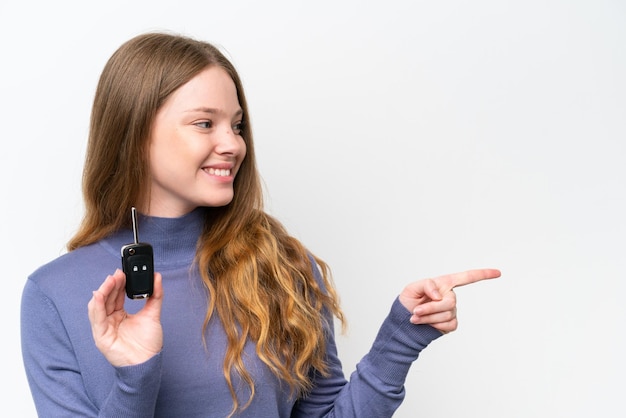 Junge kaukasische Frau, die Autoschlüssel lokalisiert auf weißem Hintergrund hält, der auf die Seite zeigt, um ein Produkt zu präsentieren