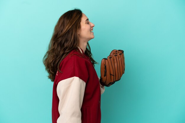 Foto junge kaukasische frau, die baseball isoliert auf blauem hintergrund spielt und in seitenlage lacht