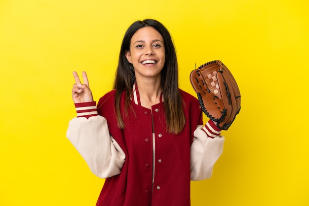 Junge kaukasische Frau, die Baseball spielt, isoliert auf gelbem Hintergrund, lächelt und zeigt Victory-Zeichen