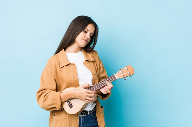 Junge kaukasische Frau, die das ukelele lokalisiert auf einer blauen Wand spielt