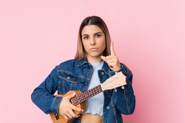 Junge kaukasische Frau, die das ukelele zeigt Nummer Eins mit dem Finger spielt.