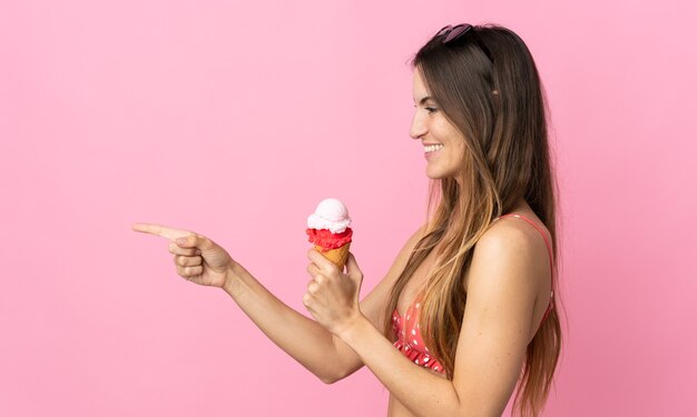 Junge kaukasische Frau, die ein Eis hält, das auf rosa Hintergrund isoliert ist und auf die Seite zeigt, um ein Produkt zu präsentieren?