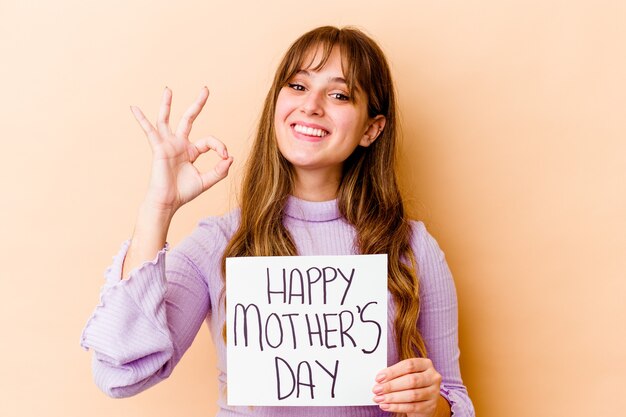 Junge kaukasische Frau, die ein glückliches Muttertagsplakat fröhlich und zuversichtlich hält, das ok Geste zeigt.