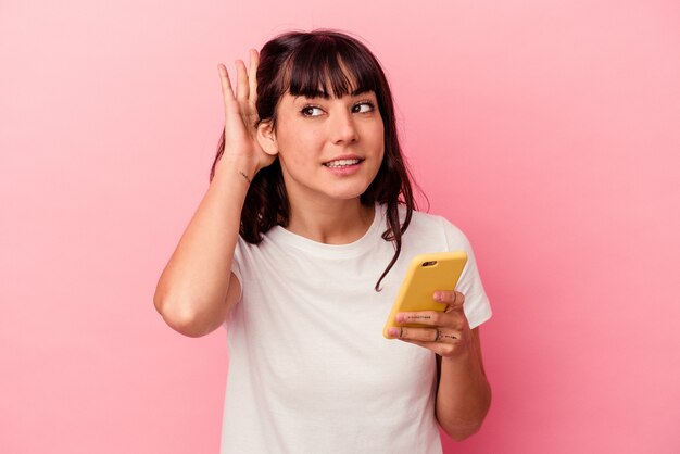 Junge kaukasische Frau, die ein Handy lokalisiert auf rosa Hintergrund hält, der versucht, einen Klatsch zu hören.