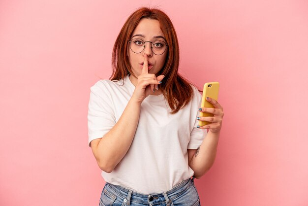 Foto junge kaukasische frau, die ein mobiltelefon isoliert auf rosafarbenem hintergrund hält, ein geheimnis hält oder um stille bittet.