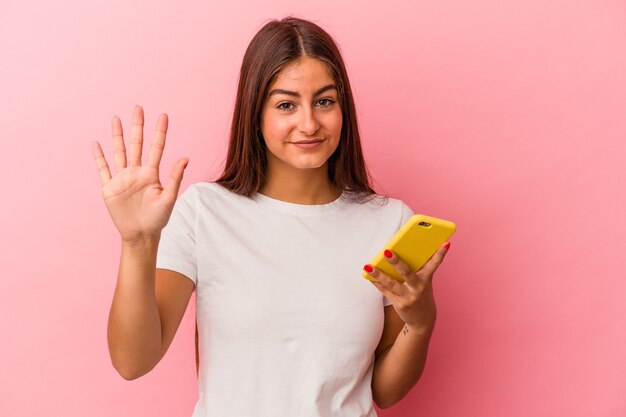 Junge kaukasische Frau, die ein Mobiltelefon isoliert auf rosafarbenem Hintergrund hält, lächelt fröhlich und zeigt Nummer fünf mit den Fingern.