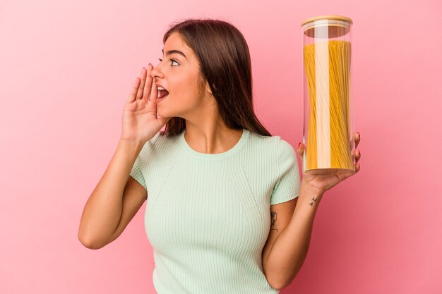 Junge kaukasische Frau, die ein Nudelglas hält, das auf rosafarbenem Hintergrund isoliert ist, schreit und Palme in der Nähe des geöffneten Mundes hält.